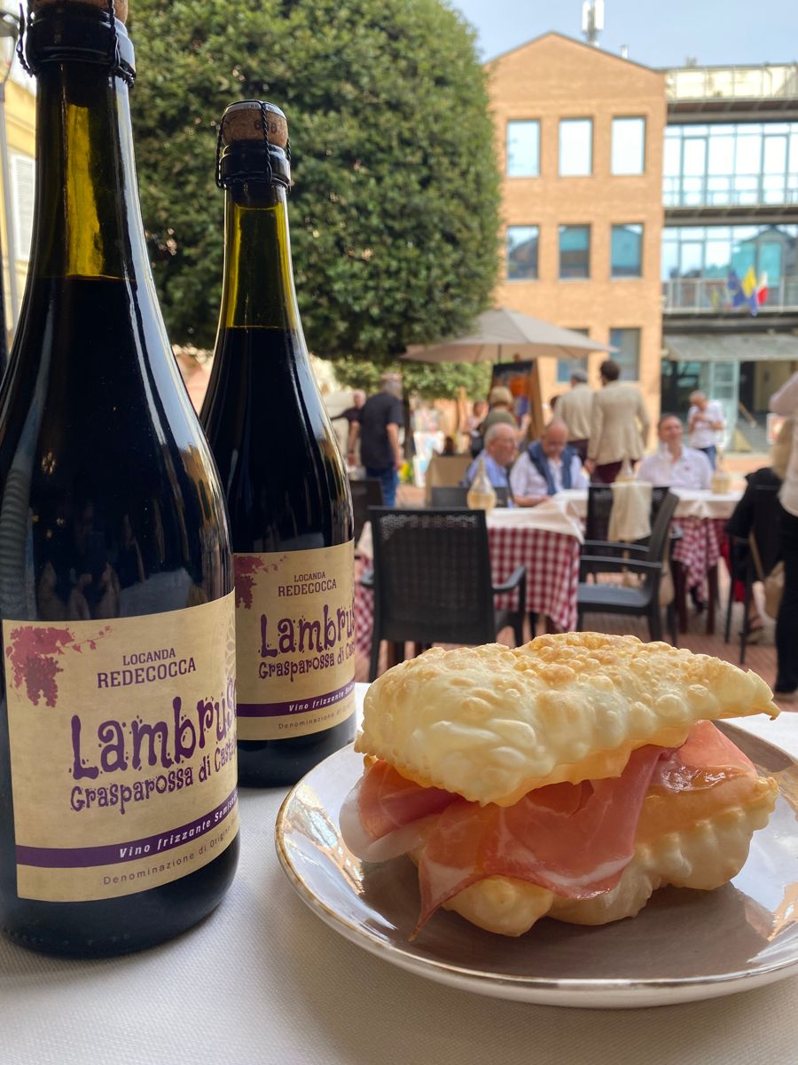 Bottiglie di Lambrusco Grasparossa con l'etichetta della Locanda Redecocca, accanto a gnocco fritto e prosciutto, ai tavoli nella piazzetta